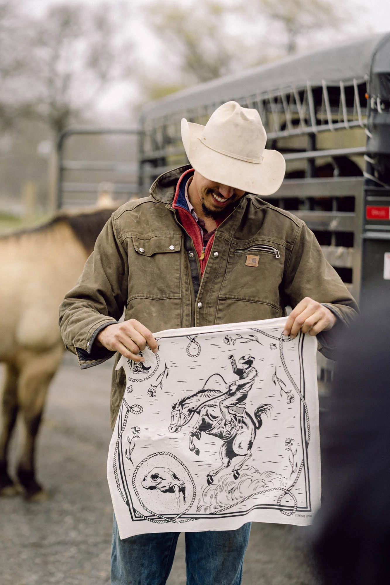 Smiling cowboy in a felt hat holding a hand-drawn bandana beside his horse