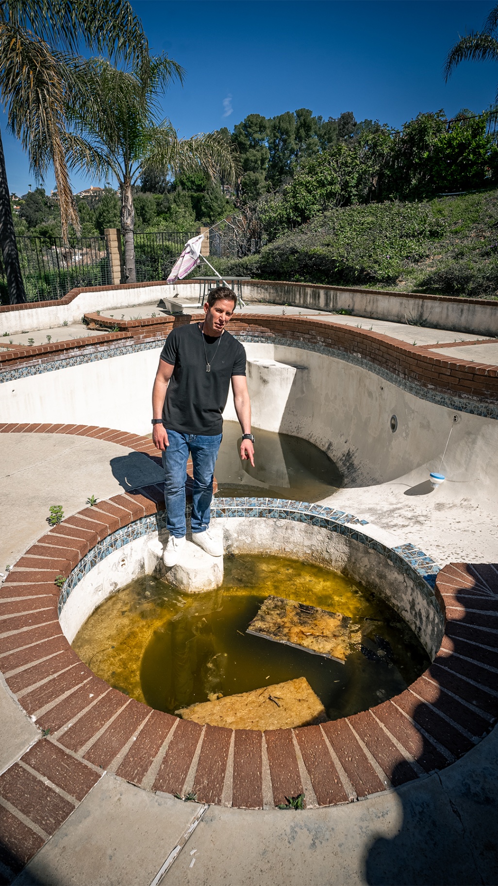 Renovation host standing beside a neglected pool with palm trees overhead